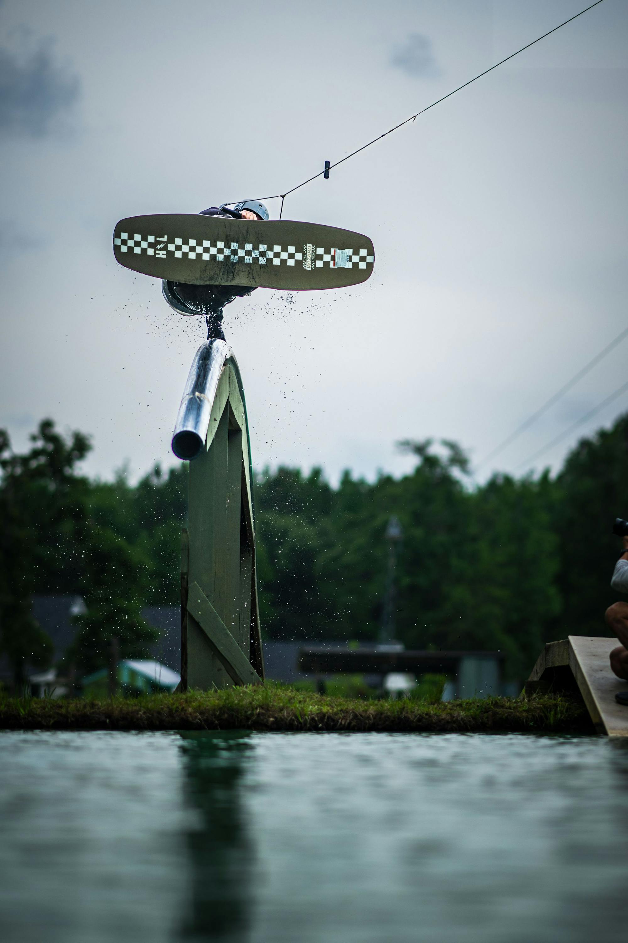 A person on a Hyperlite 2025 Wizardstick Wakeboard, likely an enthusiast of Hyperlite gear, performs an aerial trick over a metal ramp on a cloudy day.