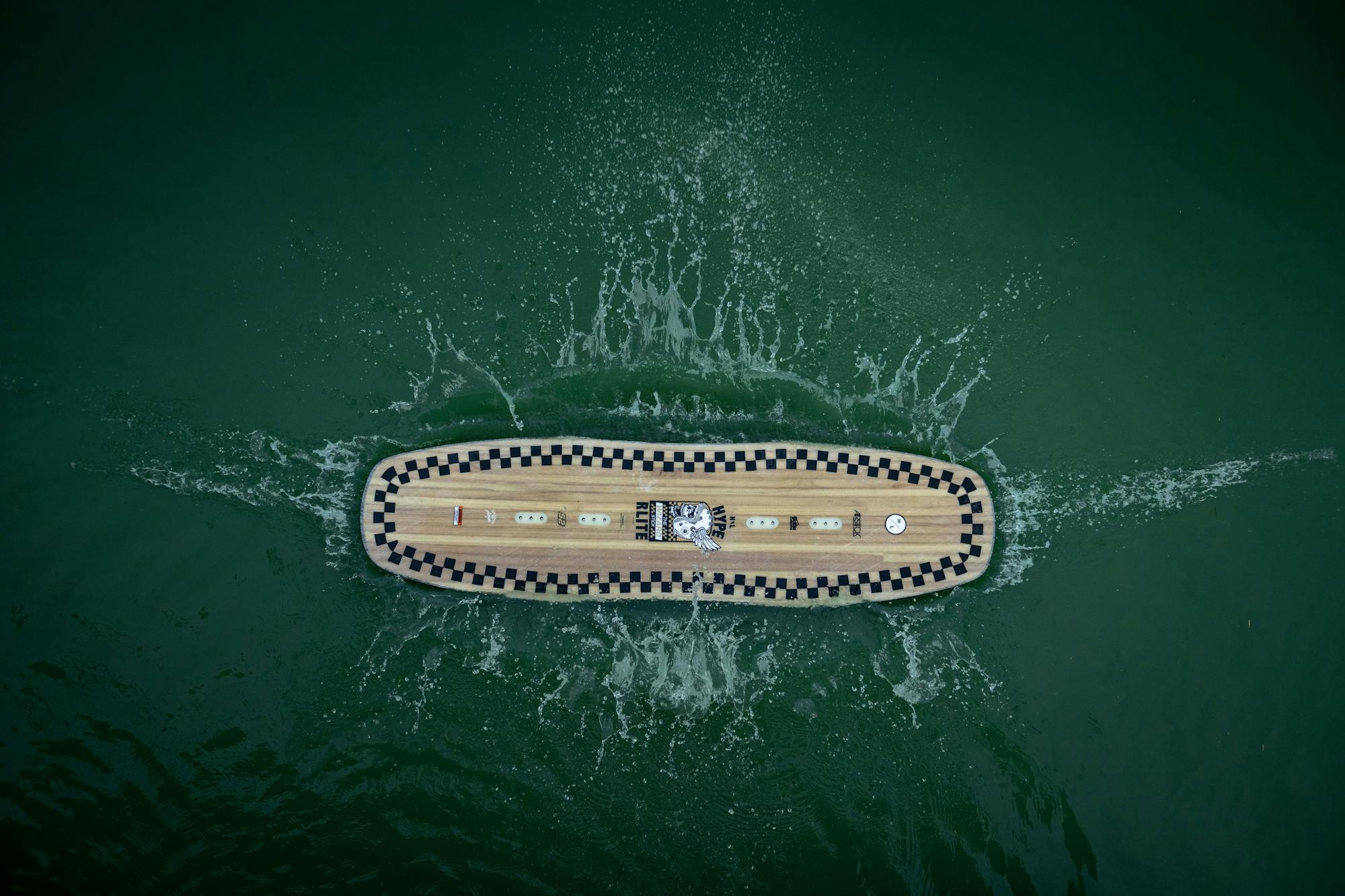 Aerial view of a Hyperlite 2025 Wizardstick wakeboard with checkered edges floating on green water, reminiscent of Trever Maur's dynamic style.