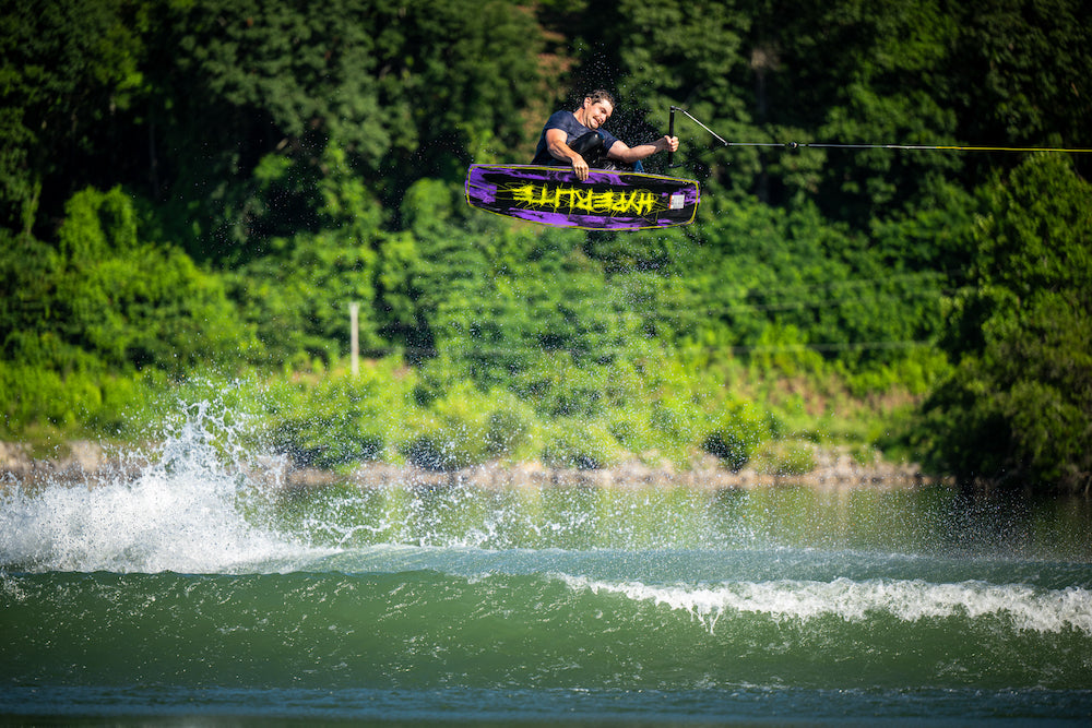 Person wakeboarding in mid-air over water with greenery in the background