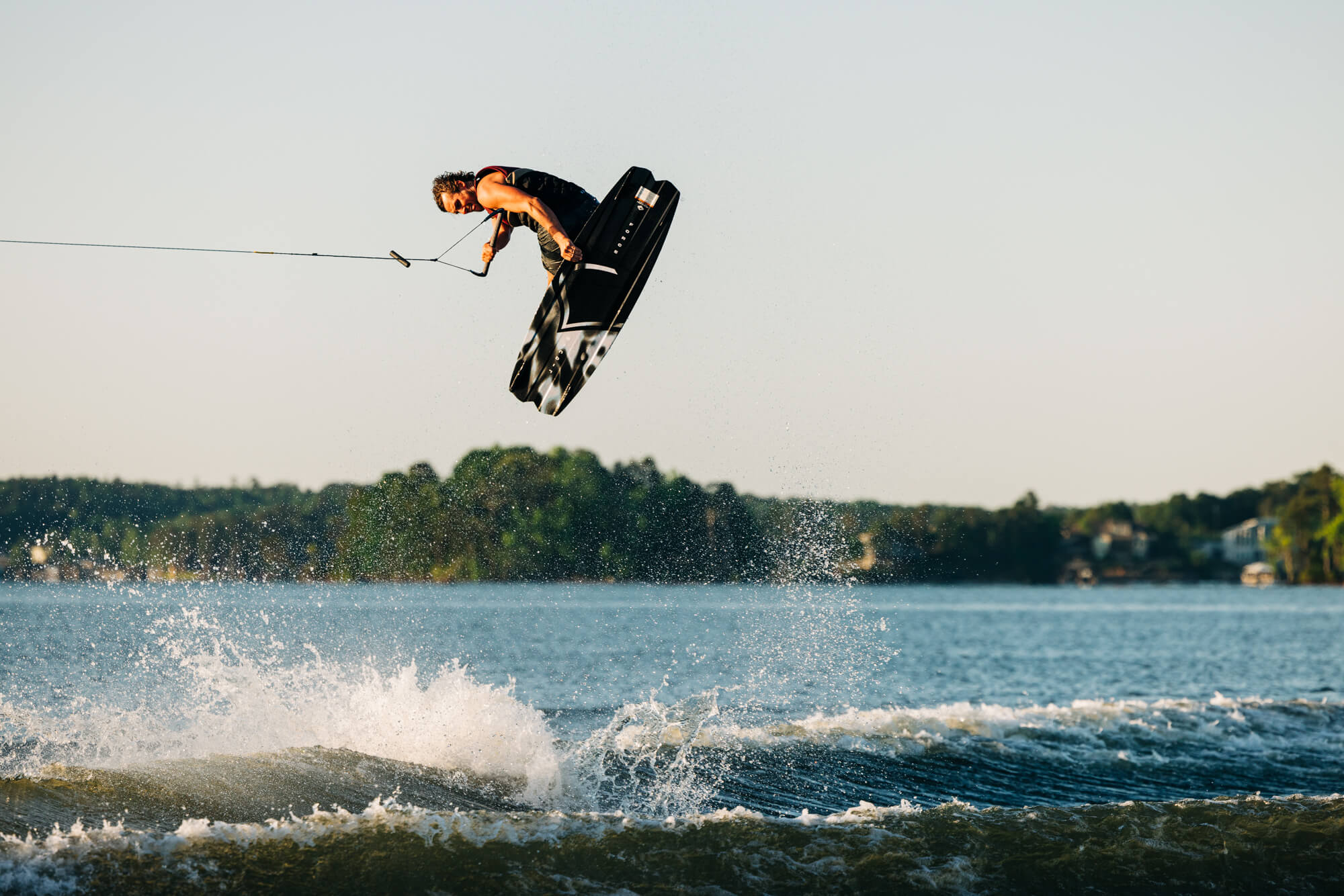Harley Clifford, a lightweight wakeboarding pro, defies gravity as he effortlessly glides through the air over a serene body of water on the Liquid Force 2024 Remedy Aero Wakeboard (Pre-Order).