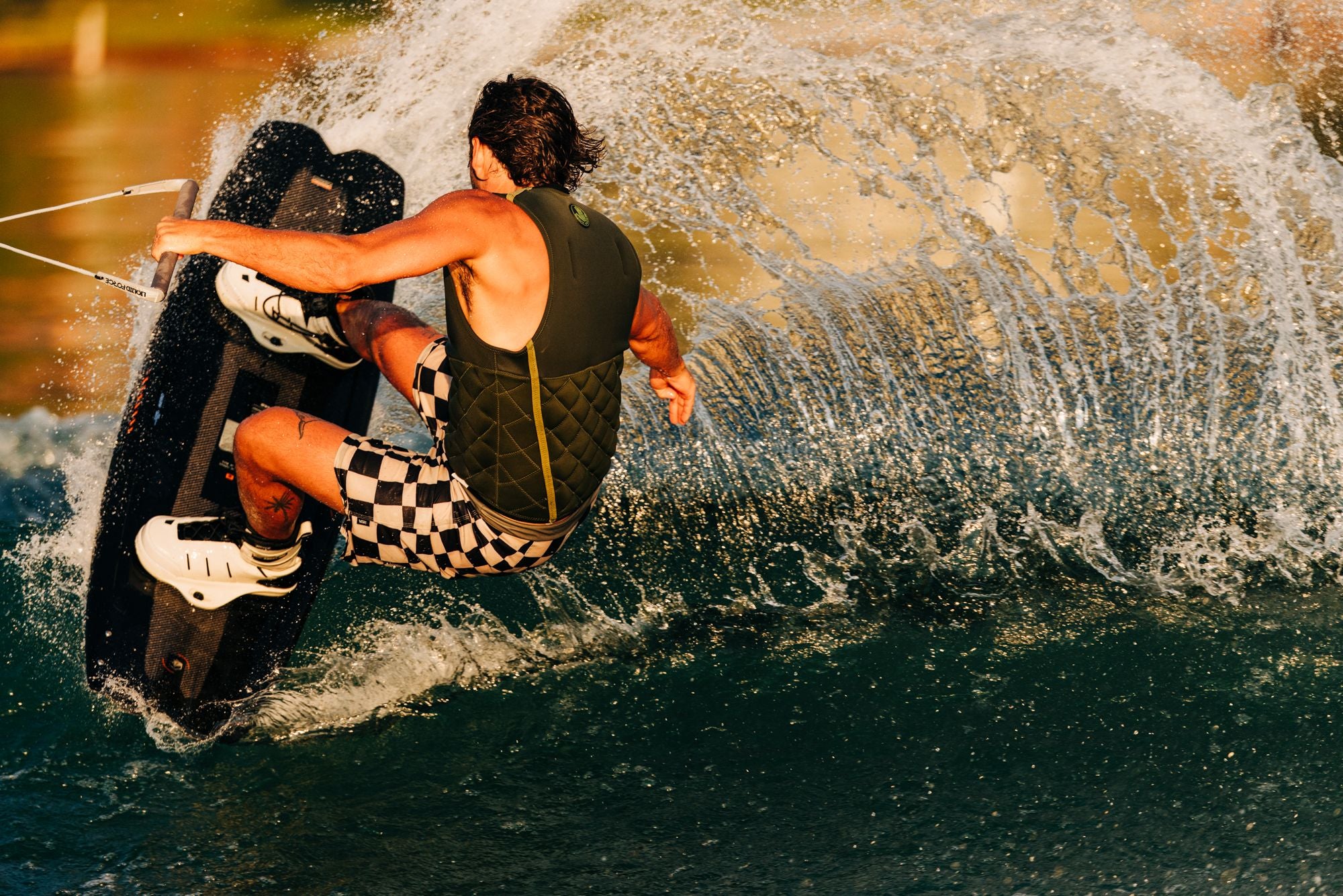 A person wakeboarding on water, wearing a life vest and checkered shorts, is equipped with Liquid Force 2025 Aero 6X Boots. The spray created by the board adds to the excitement and showcases these premium wakeboarding boots from Liquid Force.