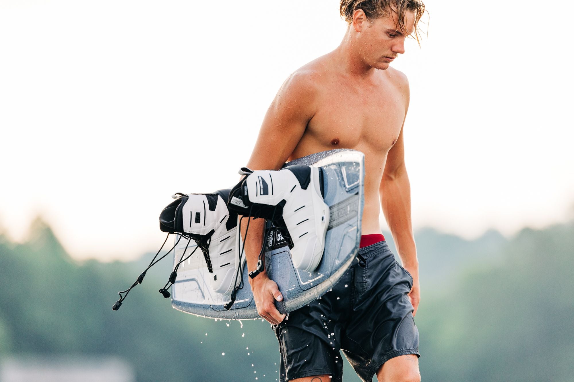 A shirtless person holding a wakeboard equipped with Liquid Force 2025 Aero 6X Boots is walking out of the water.