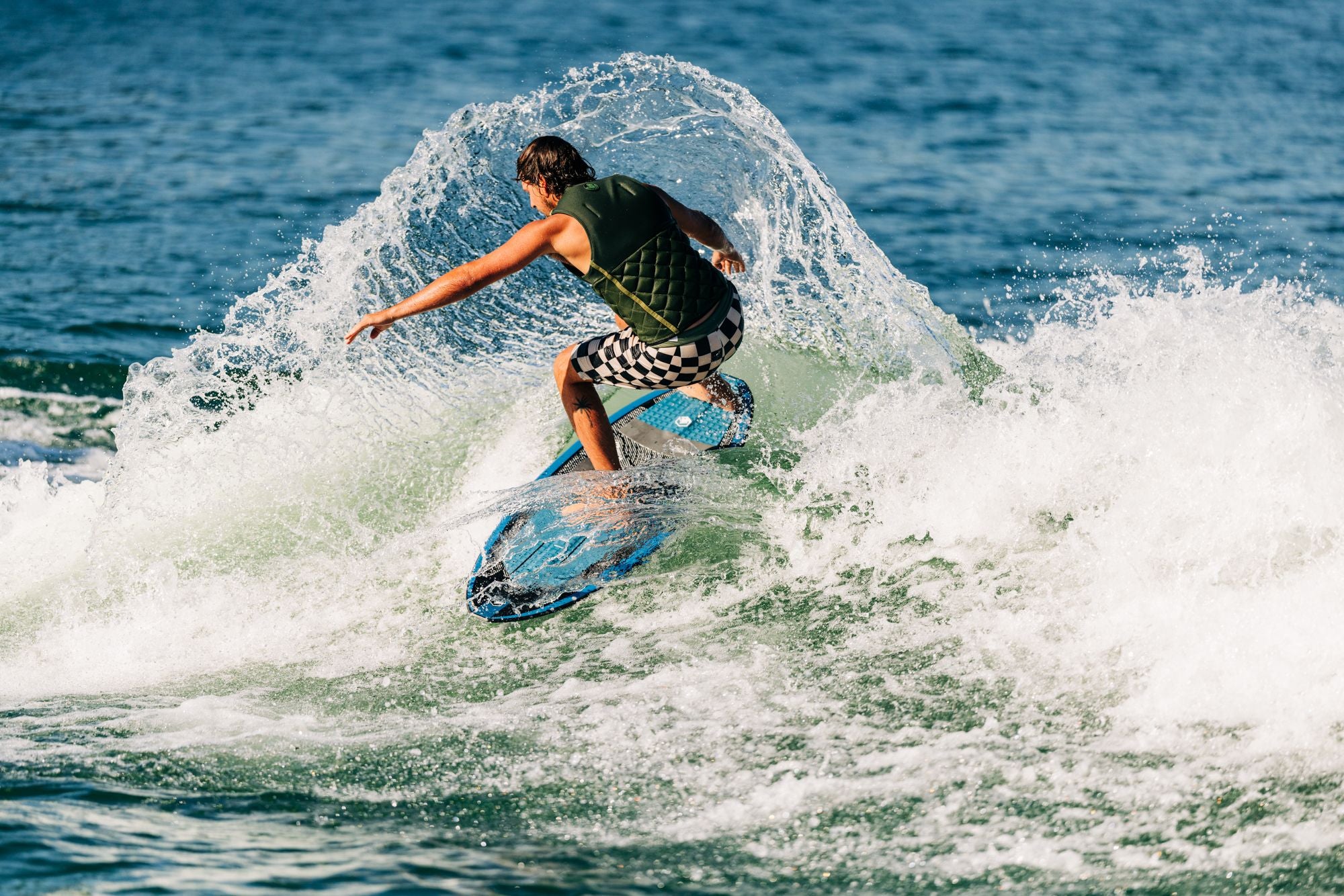 A person glides effortlessly on a Liquid Force 2025 Blade Wakesurf Board, wearing checkered shorts and a vest, against the backdrop of blue ocean water.