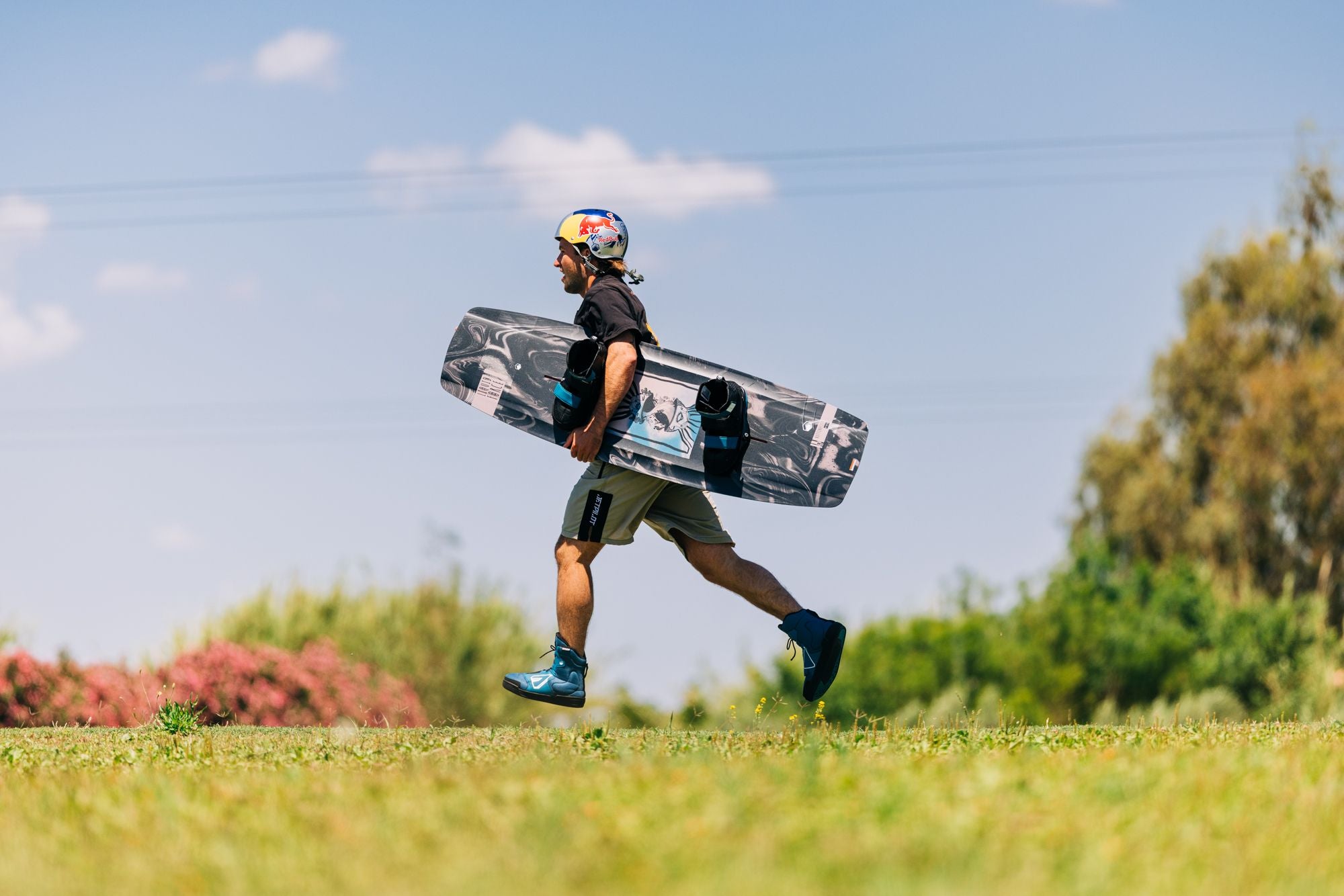 A person strolls across the grass on a sunny day, effortlessly carrying their Liquid Force 2025 Butterstick Pro Wakeboard, ready for an adventure.