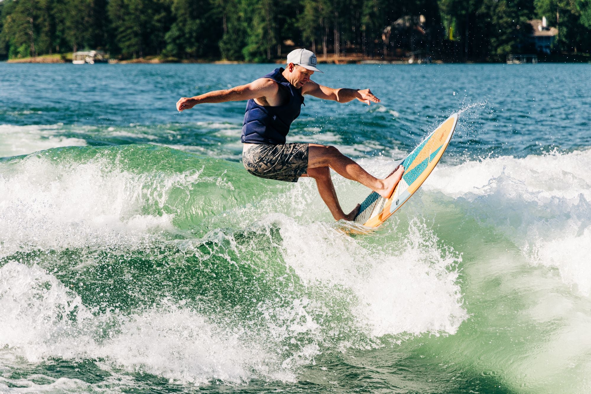 A person wearing a life vest and cap is surfing on the Liquid Force 2025 El Guapo Wakesurf Board, featuring an adjustable fin box, gracefully gliding on a wave in the tree-lined lake.