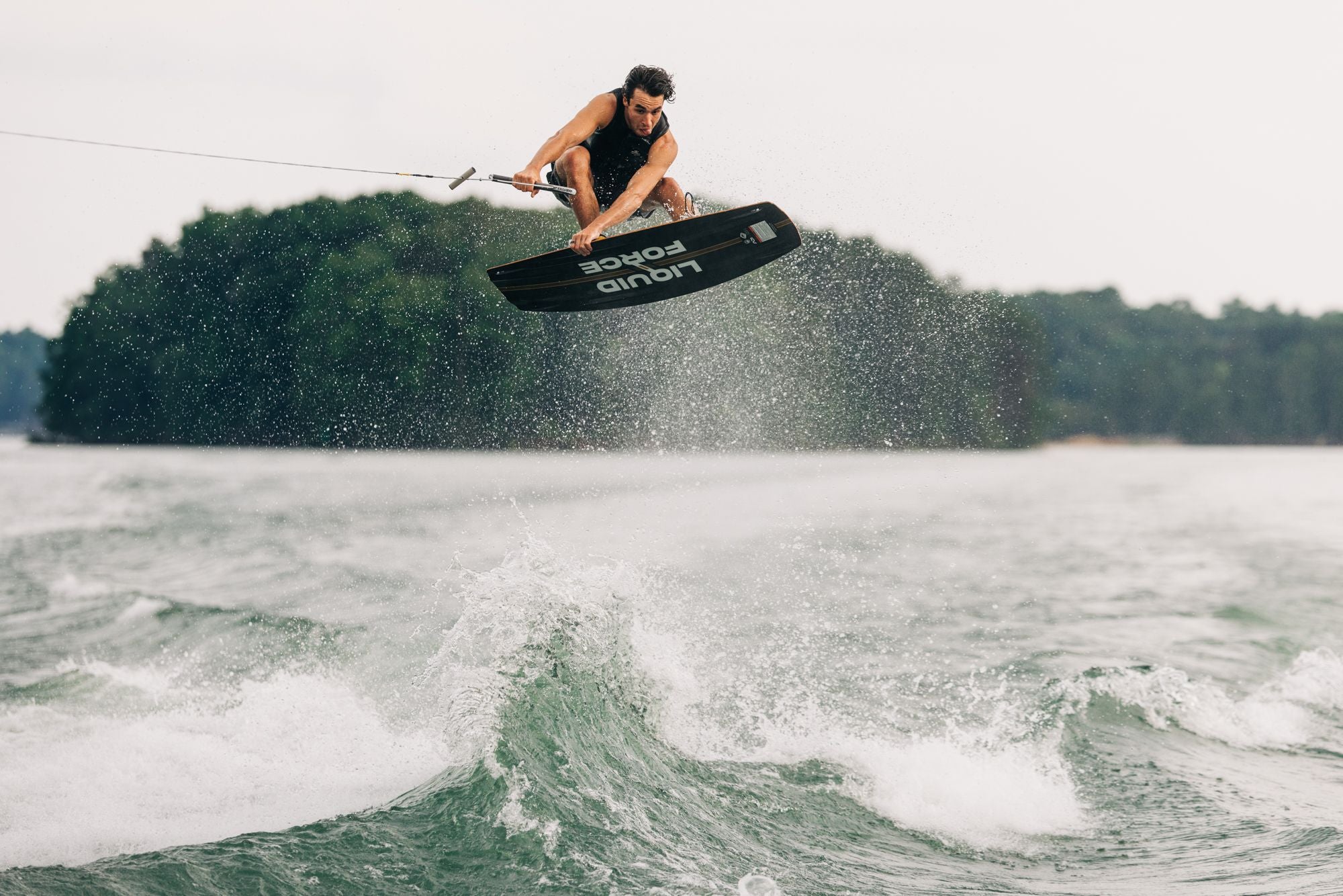 Fueled by the innovation of Liquid Force, an athlete catches air on the 2025 Ethos Wakeboard, performing a breathtaking trick over shimmering water with lush trees providing a stunning backdrop.