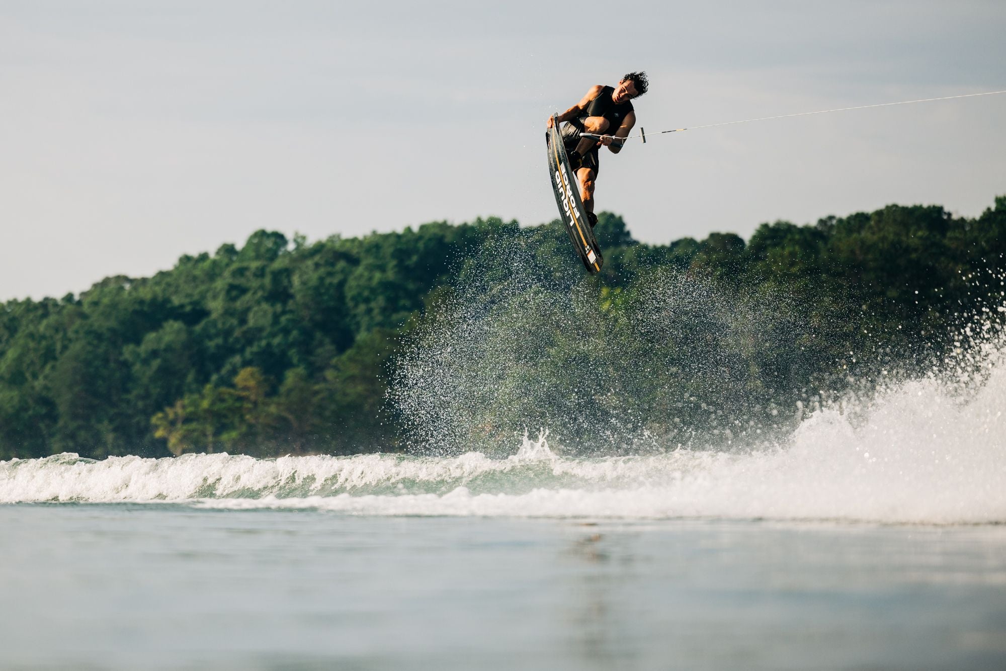 Using Liquid Force's 2025 Ethos Wakeboard, an individual showcases athlete-driven innovation in wakeboarding, soaring through the air above the water with a stunning forested shoreline as a backdrop.