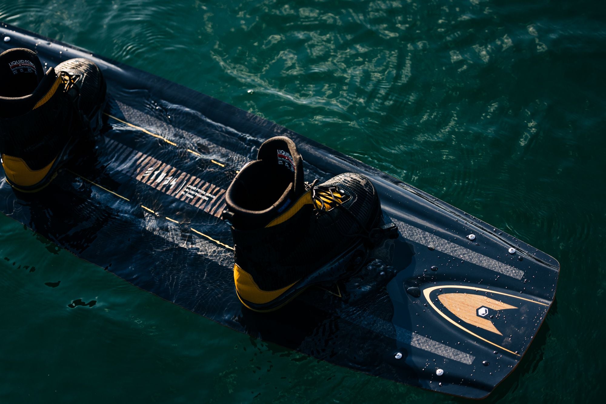 The Liquid Force 2025 Ethos Wakeboard, featuring a sleek black and yellow design and equipped with attached boots, embodies athlete-driven innovation as it floats on the water.