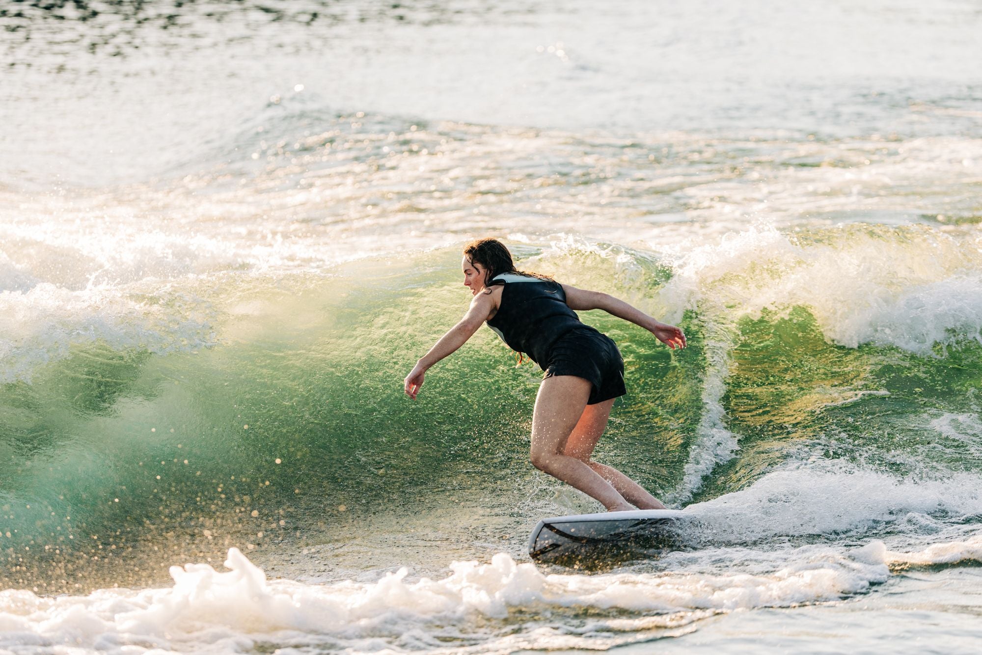 A person effortlessly carving through a green wave in the ocean, their black outfit seamlessly merging with the sleek design of the Liquid Force 2025 Flyer Wakesurf Board beneath them. The scene captures a perfect blend of thrill and glide speed as they ride with unyielding grace.