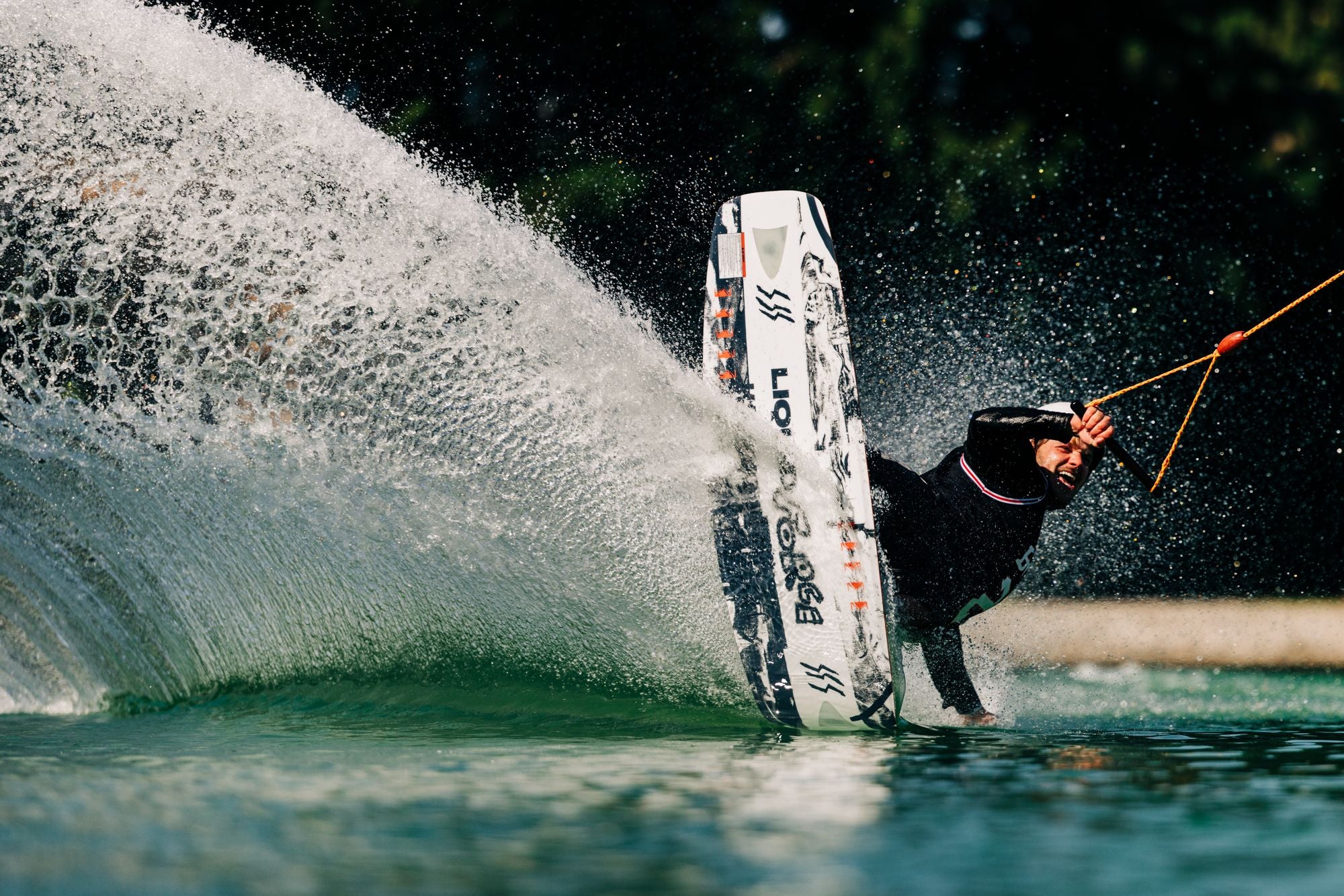 A person skillfully executes a dramatic maneuver on the Liquid Force 2025 Illusion Wakeboard, producing a large splash that blurs the line between reality and illusion.