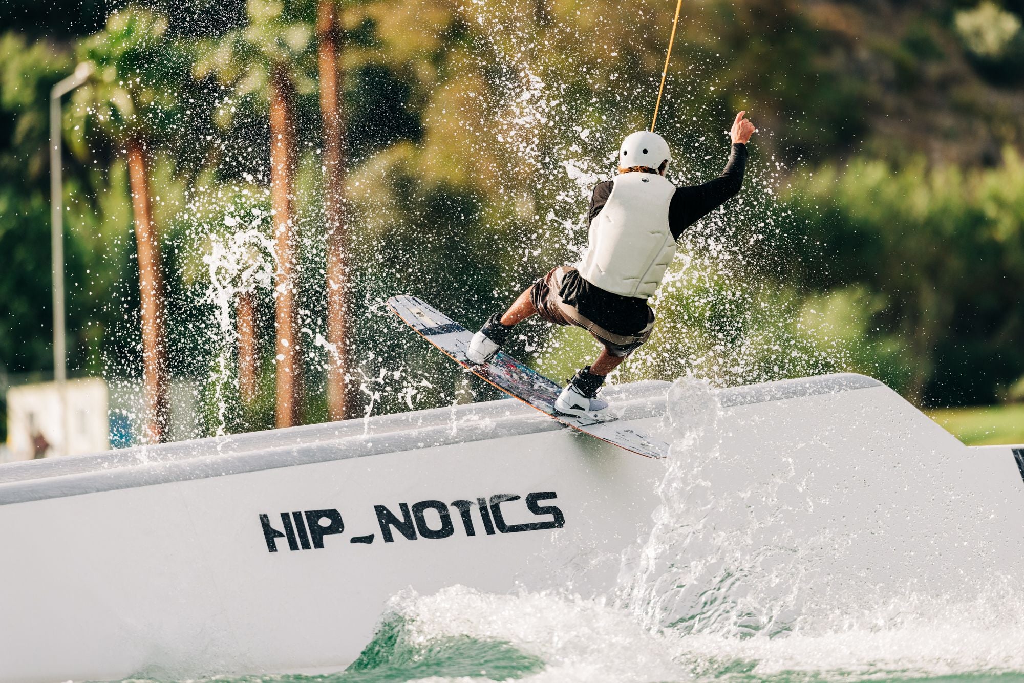 A person is wakeboarding on a ramp labeled HIP_NOTICS, showcasing the "Just The Tip" Technology of the Liquid Force 2025 Illusion Wakeboard, as they splash water while wearing a helmet and life jacket.