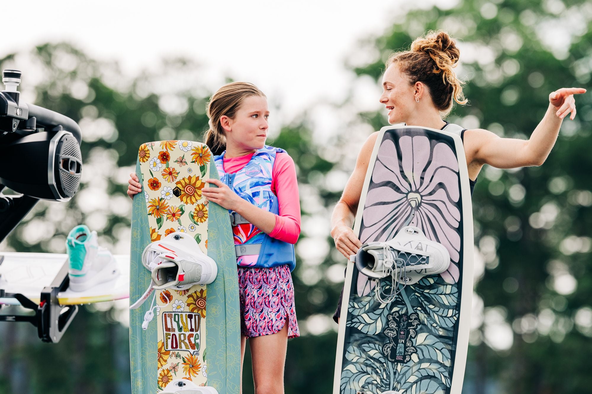 Two younger riders hold their Liquid Force 2025 M.E. Kid's Wakeboards on a dock, with one pointing into the distance.