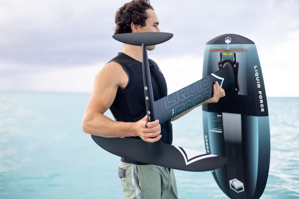 A man stands by the ocean, clutching a 2025 Liquid Force Nebula Foil Board with cutting-edge Dura-Surf Construction. He's dressed in a black vest and khaki shorts, ready to hit the waves with his trusty hydrofoil.