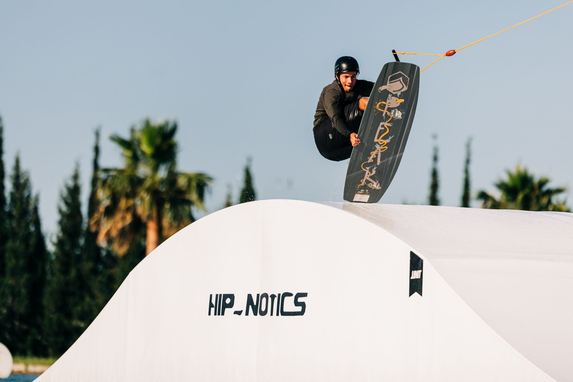 A person using the Liquid Force 2025 Peak Wakeboard, while wearing a helmet, executes a lightweight pop over a white ramp emblazoned with Hip-Notics, set against a backdrop of palm trees.