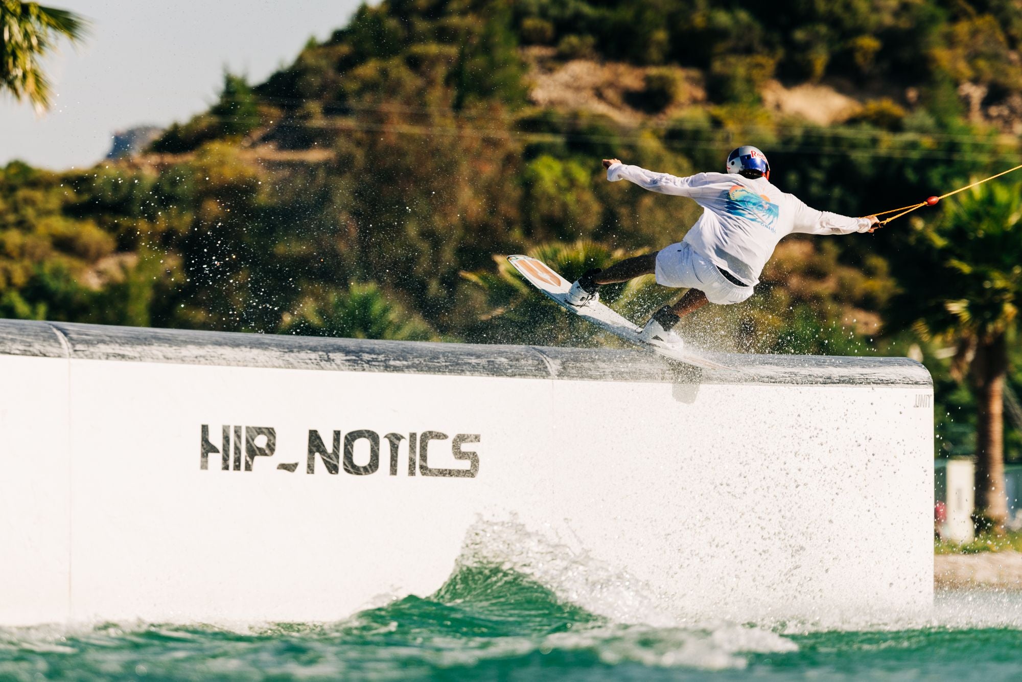 A person in a white outfit is wakeboarding on a rail at Hip-Notics, demonstrating the lightweight pop of the Liquid Force 2025 Peak Wakeboard, with trees and a hill in the background.