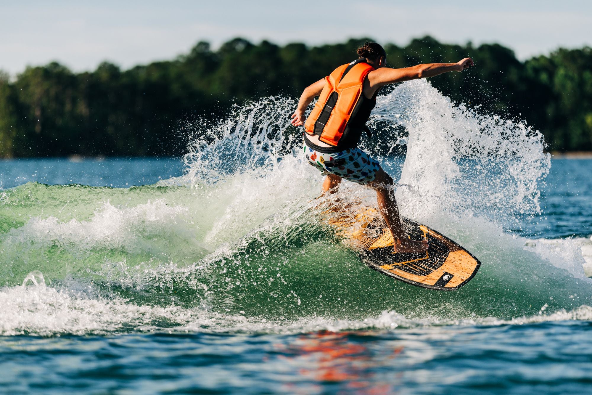 Wearing an orange life jacket, a person skillfully maneuvers on the Liquid Force 2025 Pod Wakesurf Board, slicing through the waves with precision. In the background, trees border the shore, adding a peaceful setting to the exhilarating surf adventure.