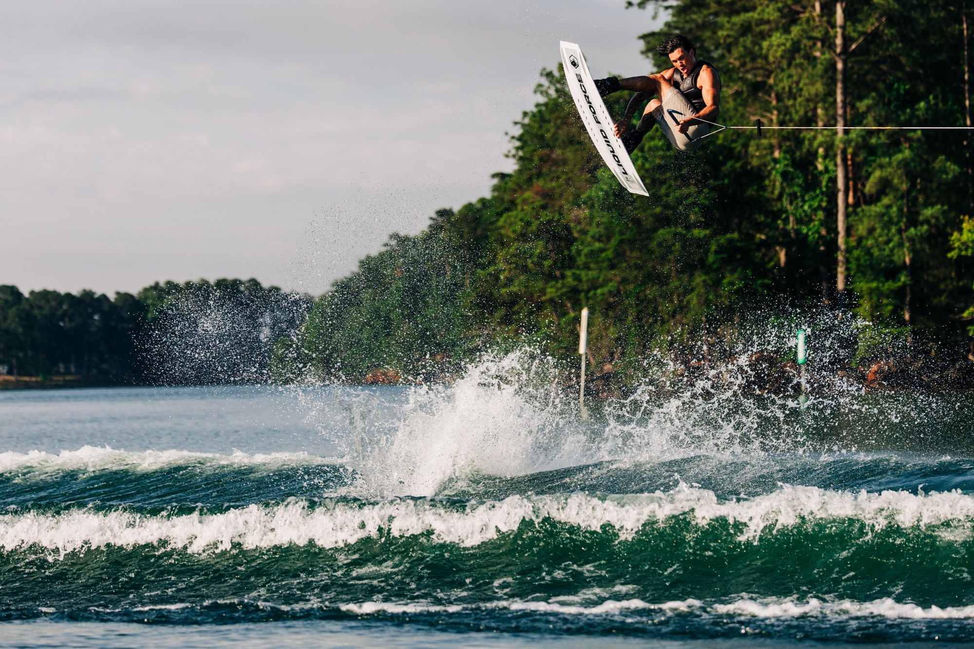 Fynn Bullock expertly handles the Liquid Force 2025 Prime Aero Wakeboard, soaring above the water in a spectacular jump, framed by a backdrop of lush trees.