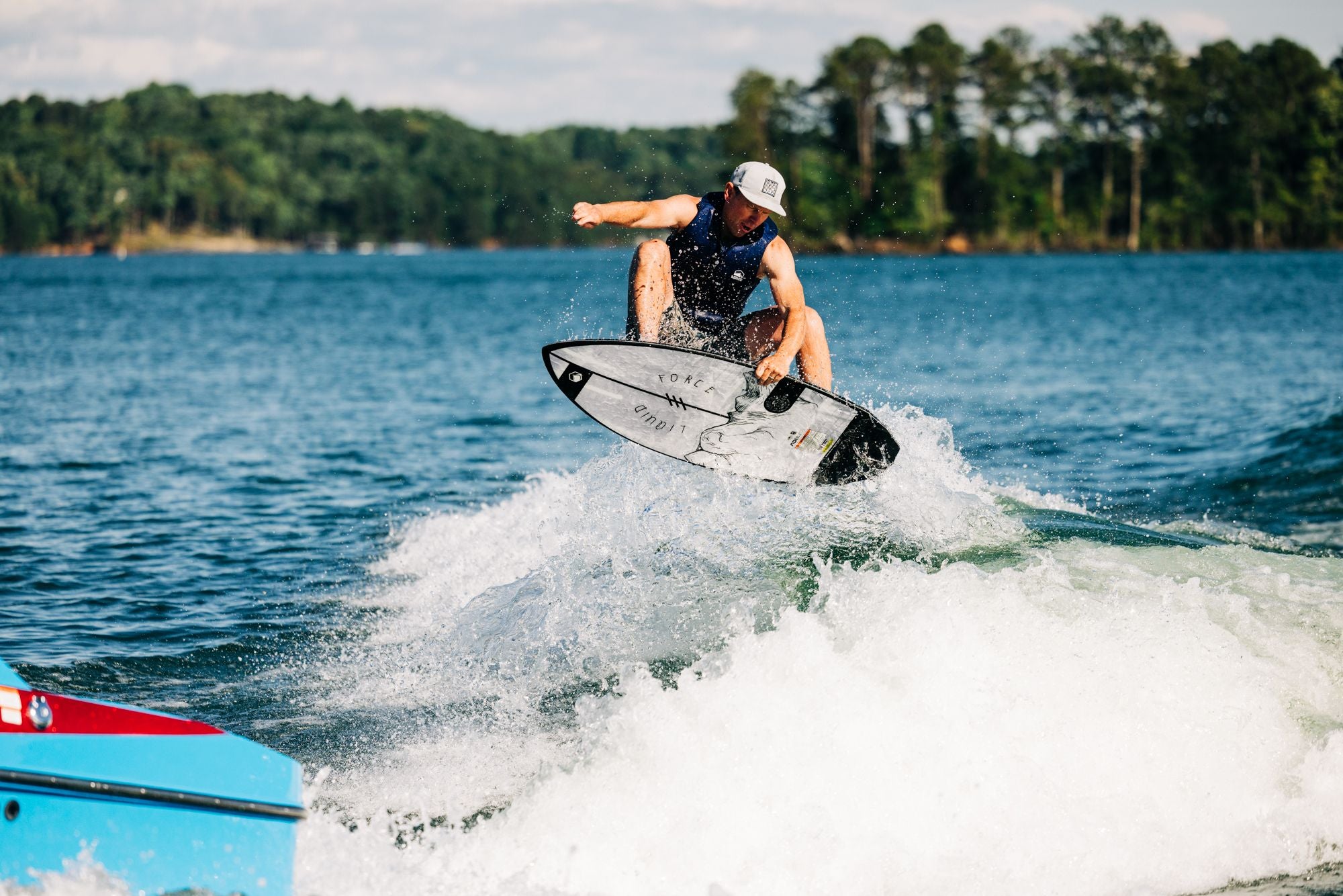 A person wakesurfs on a lake, effortlessly combining a surf-style vibe with their jump using a Liquid Force 2025 Primo Wakesurf Board. A red and blue boat is partially visible in the foreground as trees edge the distant shore beneath a clear sky.