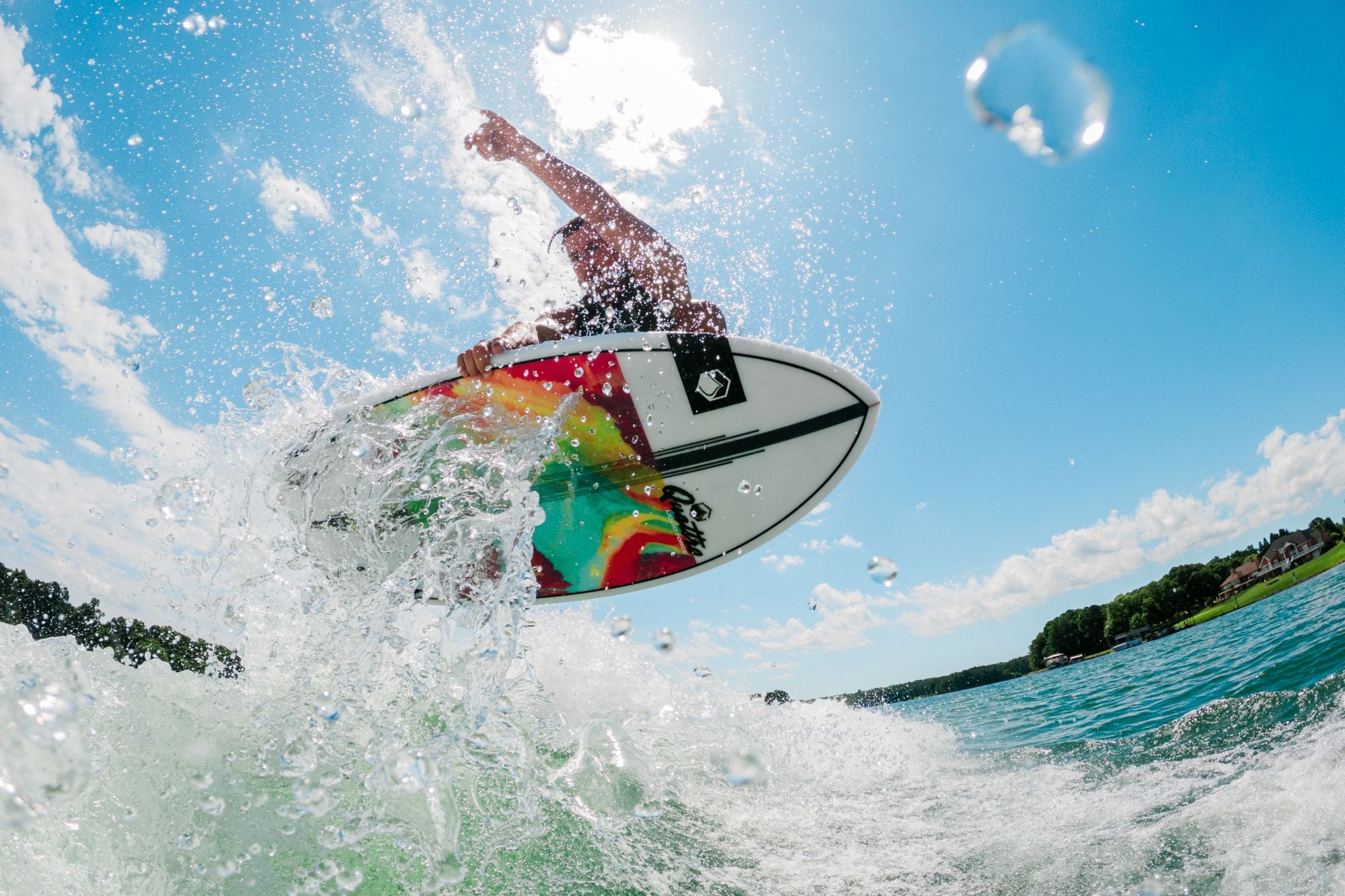 A surfer catches air on a wave, riding a vibrant Liquid Force 2025 Quattro Wakesurf Board against the sunny blue sky. Water droplets sparkle in the foreground, highlighting the surf-style artistry of their ride.