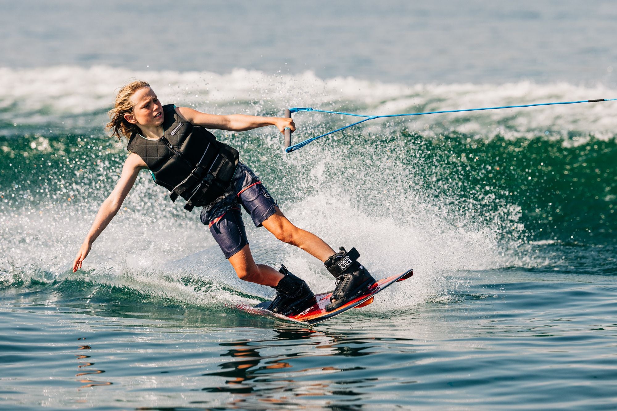 A younger rider is wakeboarding on a lake, wearing a life vest and shorts from the Liquid Force Future Series, gripping a rope handle with water splashing around, while enjoying the Liquid Force 2025 RDX Kid's Wakeboard.