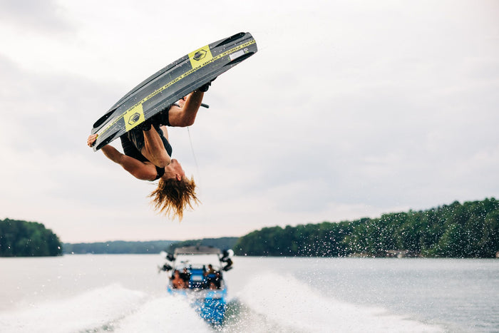 An individual executes a gravity-defying stunt on the Liquid Force 2025 RDX Wakeboard with Transit 6R Bindings, landing smoothly and effortlessly, while a boat and water form the backdrop.