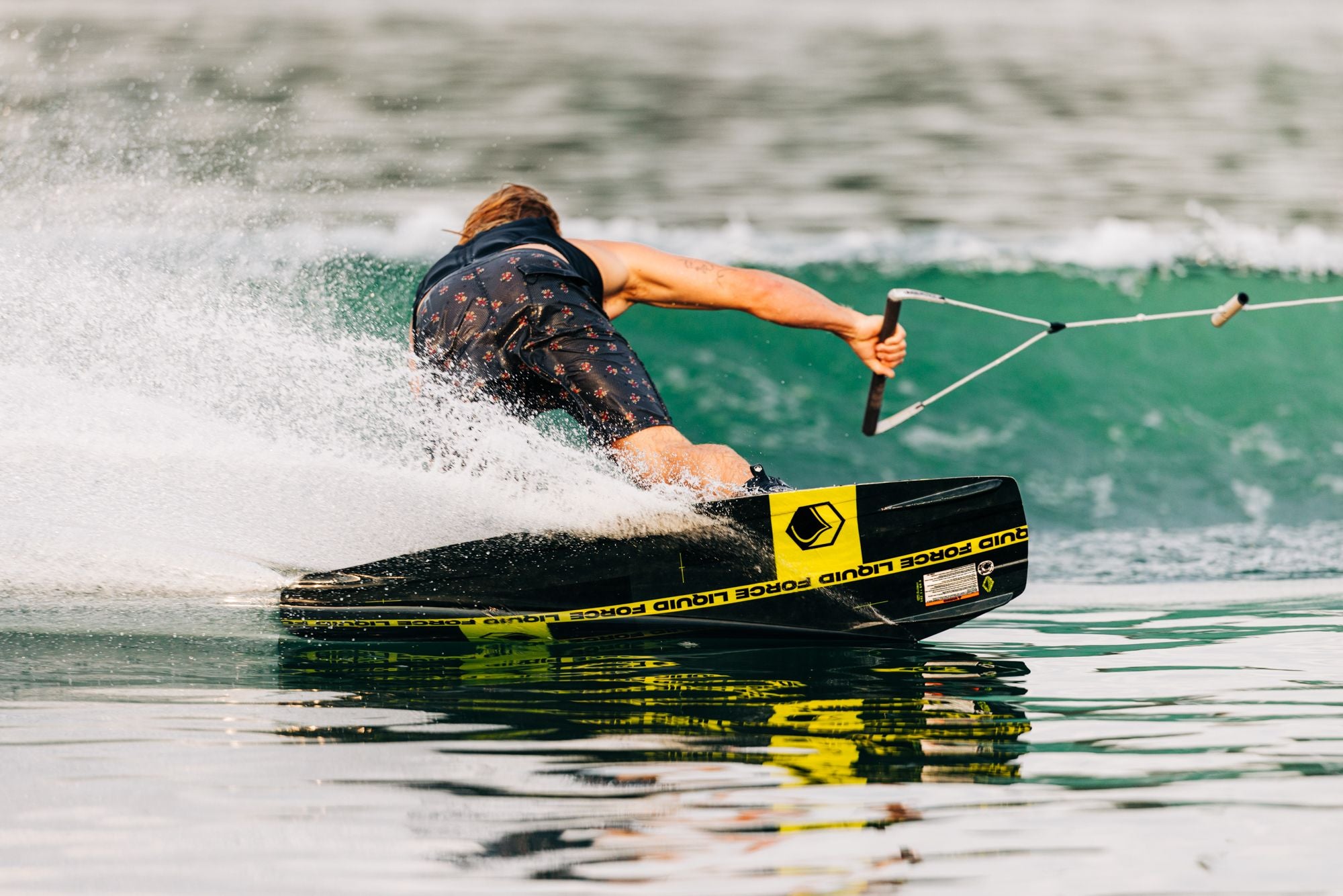 A person water skiing on a calm lake, creating a spray of water while holding onto a rope handle and gliding effortlessly on a Liquid Force 2025 RDX Wakeboard.