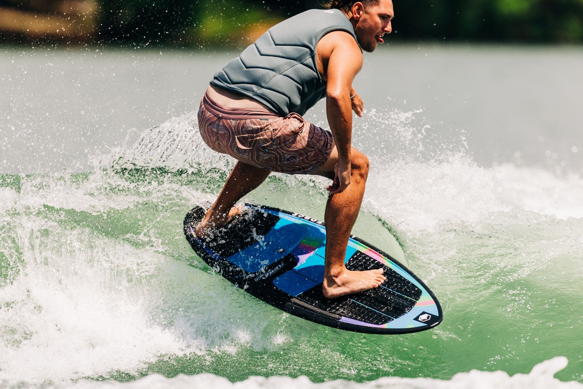 A person clad in a sleeveless vest and shorts is wakesurfing on a wave, masterfully showcasing their skills with the Liquid Force 2025 Reign Pro Wakesurf Board in a sunny outdoor setting. For more thrilling moments like this, check out Jake’s Instagram featuring the Reign Pro.