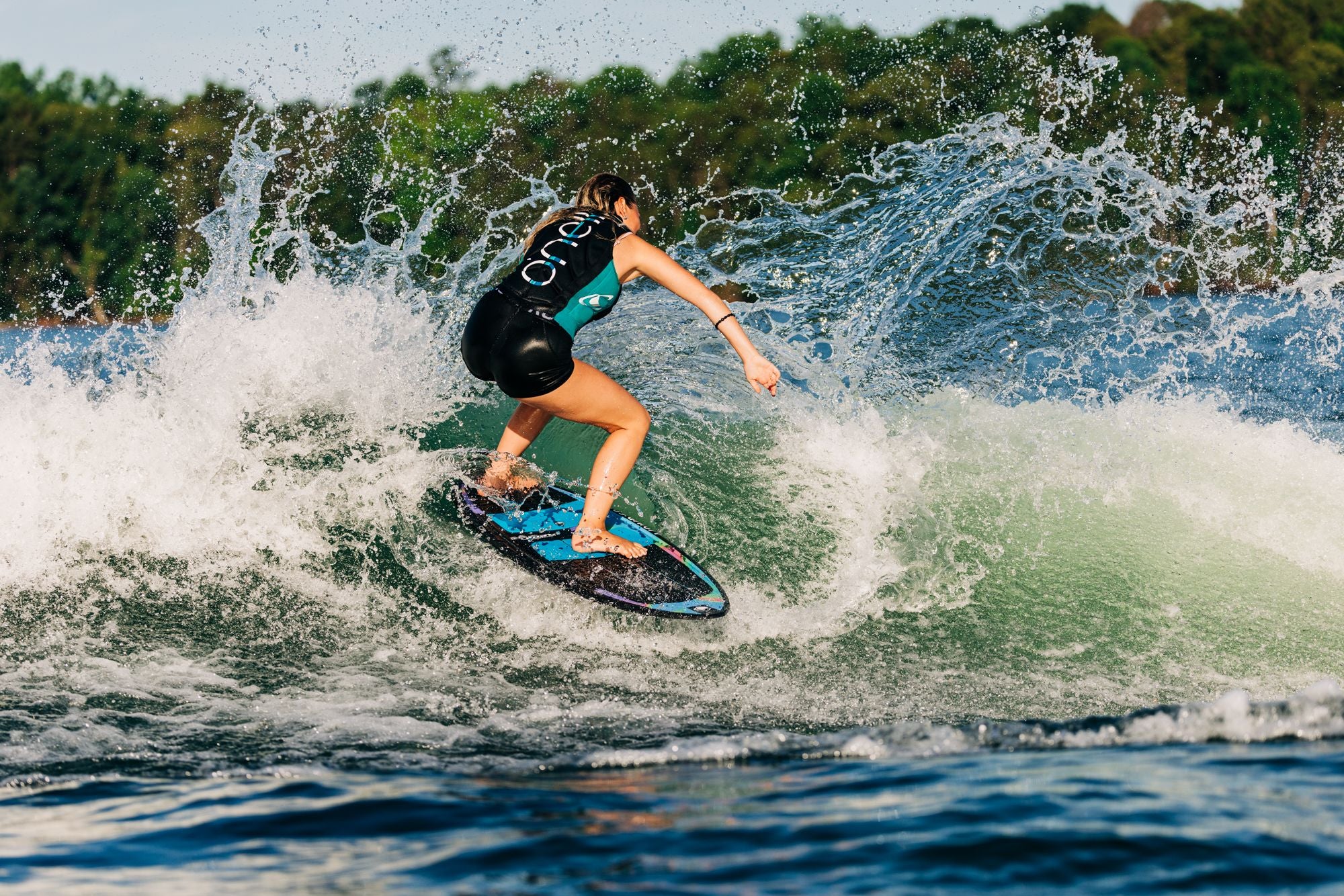 A person wakesurfing on a wave, clad in a black wetsuit and maintaining balance on a Liquid Force 2025 Reign Pro Wakesurf Board, with trees visible in the background.