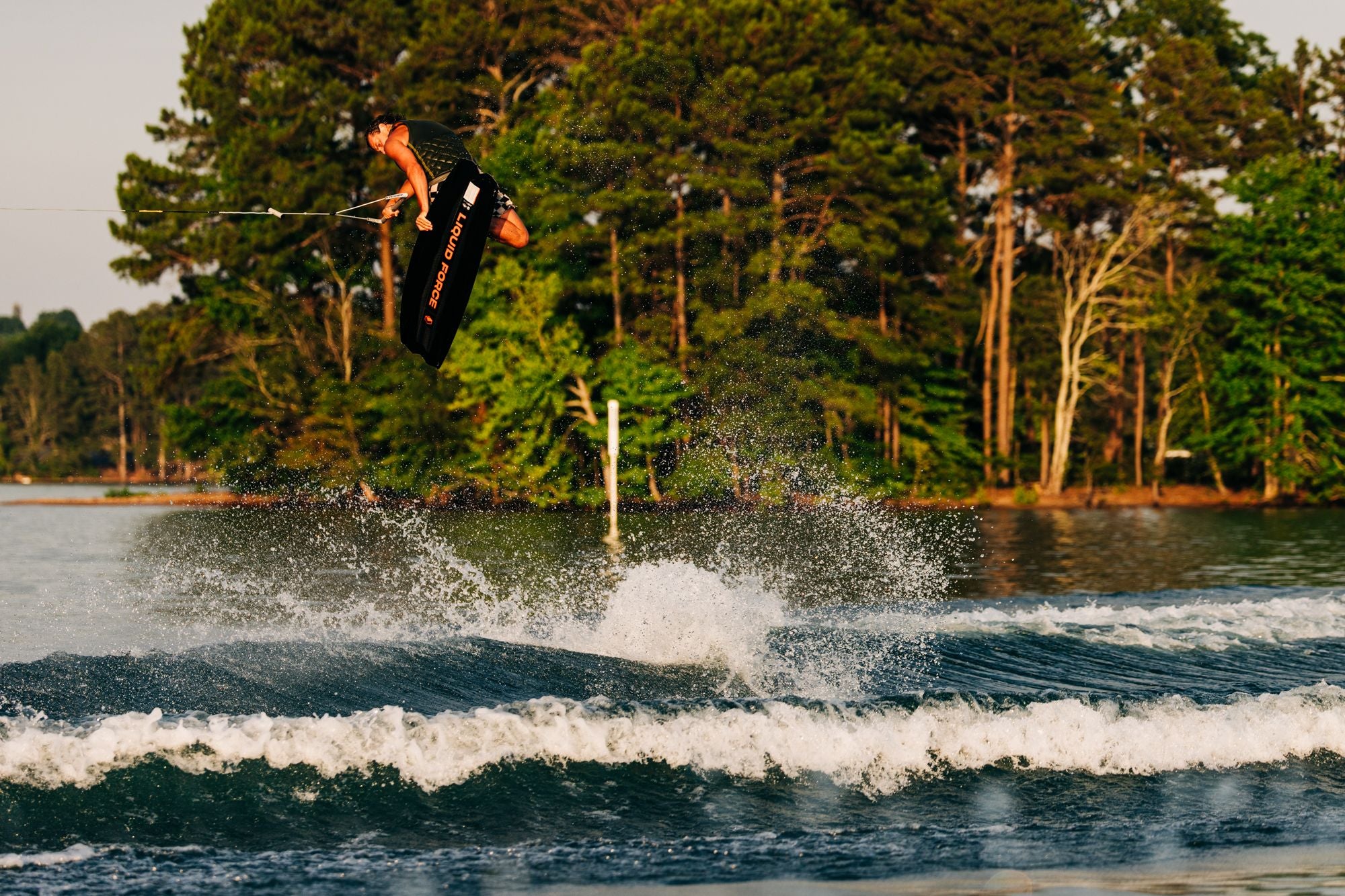 A person wakeboarding on a Liquid Force 2025 Remedy Aero Wakeboard executes an aerial trick over the water, with trees visible in the background. The sleek design of the board enhances their agility and style mid-air.