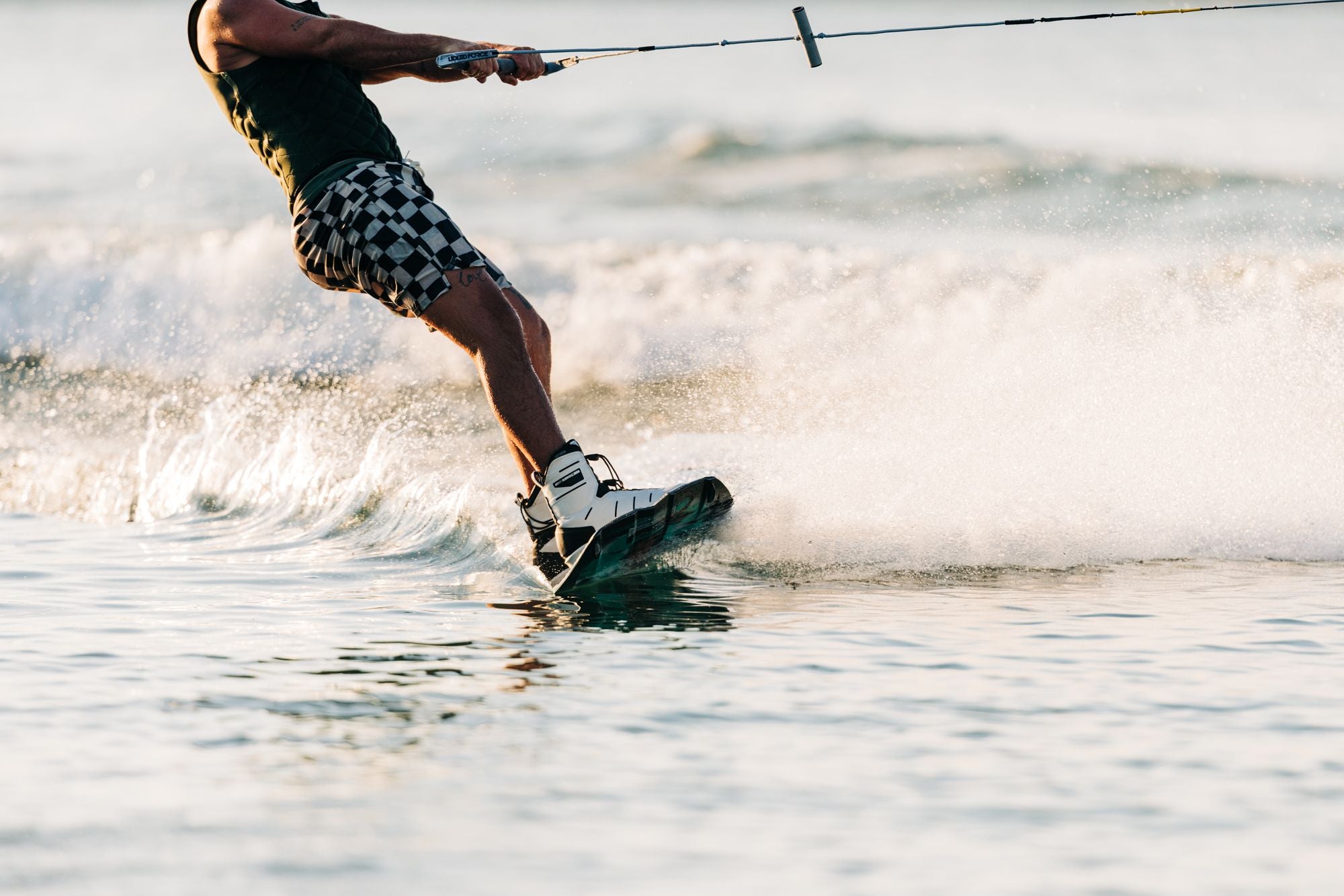 A person wakeboarding on the water, clad in checkered shorts and a life vest, expertly gripping a tow handle for an exhilarating journey with the Liquid Force 2025 Remedy Wakeboard.