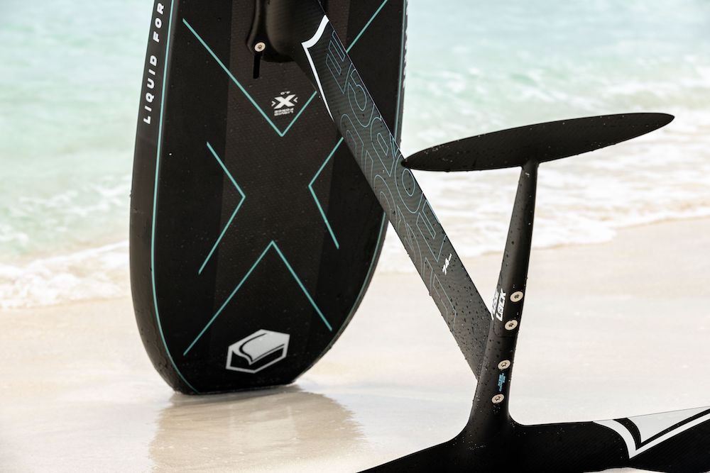 A black Liquid Force 2025 Stratus Carbon 950 Fuselock Foil Set surfboard with blue accents, showcasing the high-performance front wing, stands on the sandy beach near the water's edge.