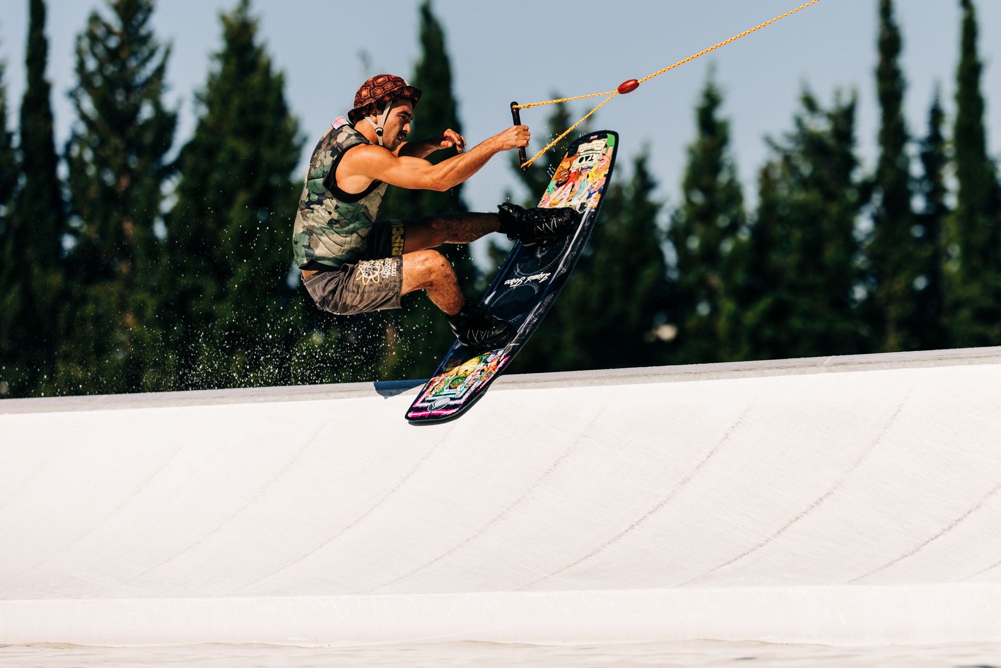 A person wakeskating on the Liquid Force 2025 Tao Wakeboard, wearing a helmet and life vest, holding a rope with one hand, set against a backdrop of trees.
