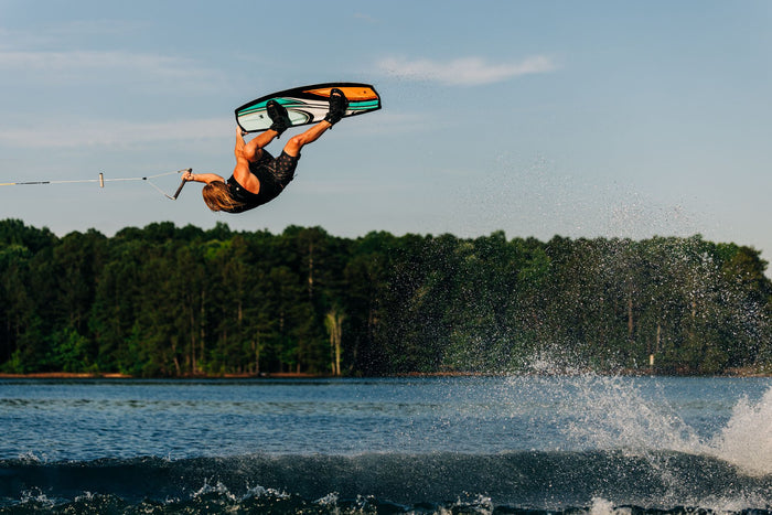 A person is wakeboarding over a lake on the Liquid Force 2025 Trip Wakeboard with Index 6R Bindings, executing an aerial trick with a sense of fun against a backdrop of trees and blue sky.