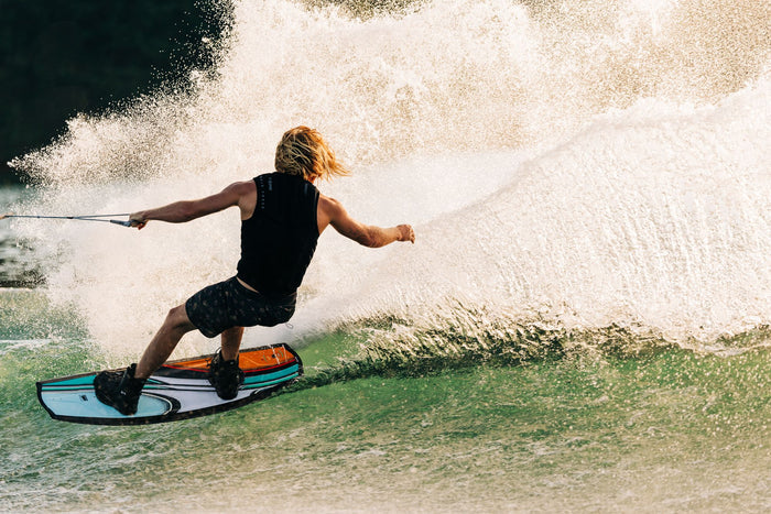 A person with long hair is wakeboarding on a wave, enjoying the sun while wearing a black life vest and shorts, skillfully holding a rope with an outstretched arm on their Liquid Force 2025 Trip Wakeboard equipped with Index 6R Bindings from Liquid Force.