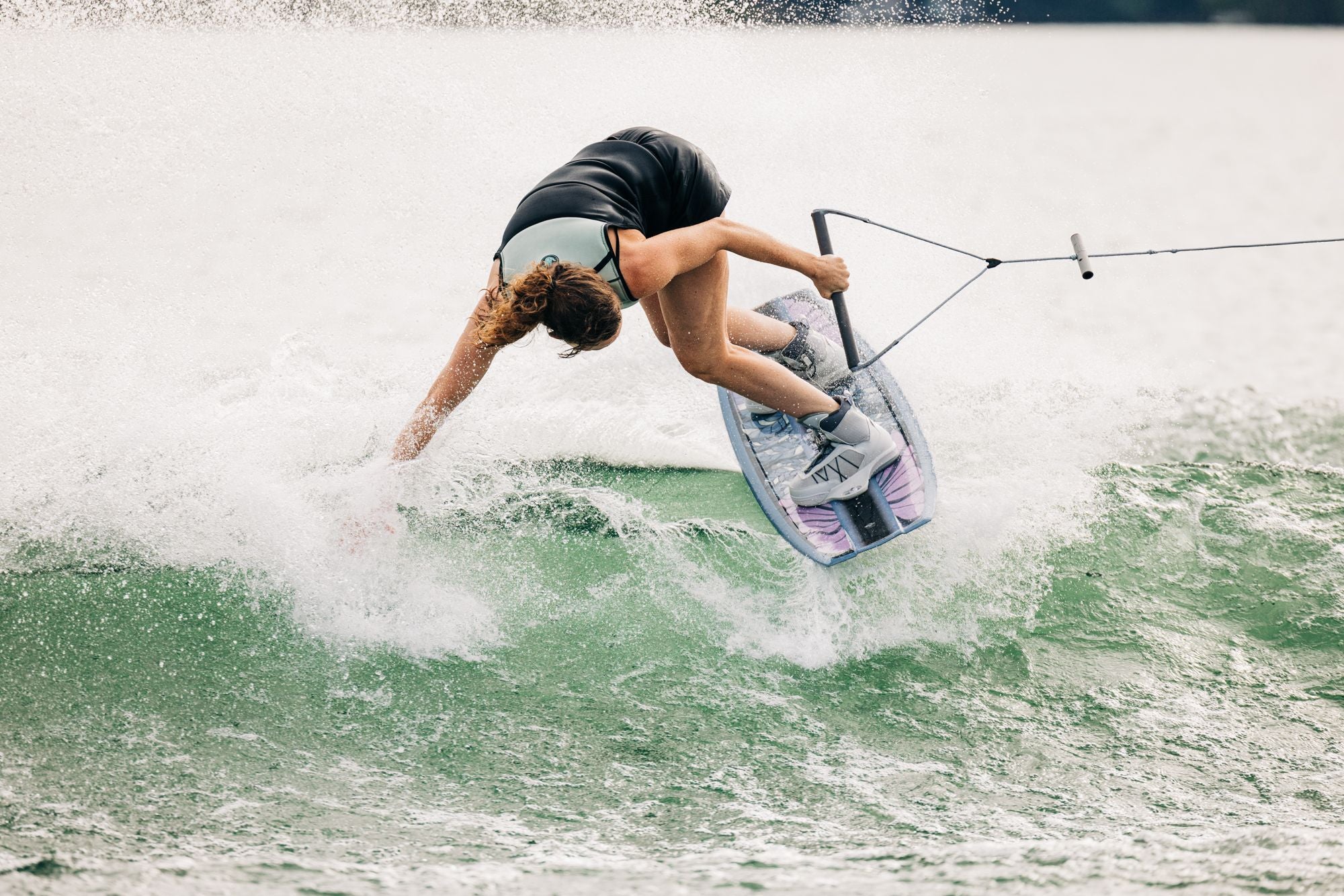 A person expertly wakeboarding on a lake performs a trick with precision, holding onto the rope handle as water splashes around them. Their movements are expertly controlled, thanks to the unparalleled support and agility provided by the Liquid Force 2025 Vida 6X Bindings.