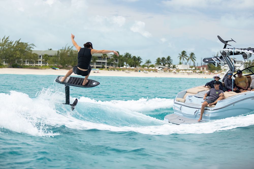 A person navigates the Liquid Force 2025 X Foil Board, showcasing its sleek, surf-inspired design on the ocean near a boat, captivating onlookers. Beach and palm trees frame the scene as wake foil aerials enhance their exhilarating ride.