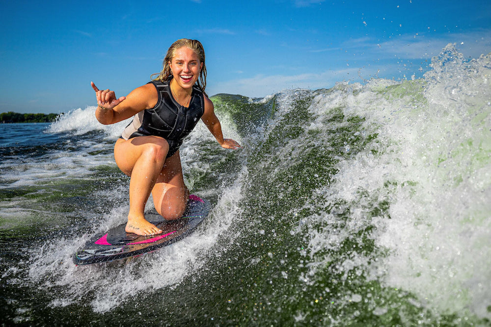 On a sunny day, a person surfs a wave with one hand raised in delight, wearing a black life jacket and smoothly gliding across the water on their Phase 5 2025 Diamond Luv Wakesurf Board.