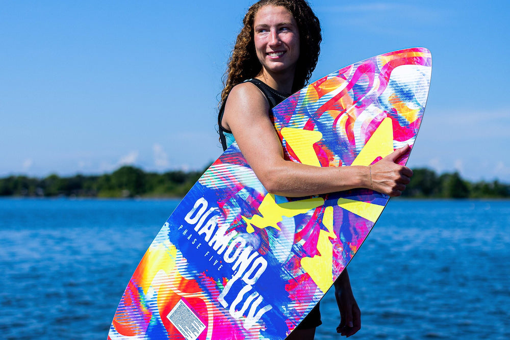 A person holding a vibrant surfboard with "Phase 5 2025 Diamond Luv Wakesurf Board" written on it stands by a body of water under a clear blue sky, their board glistening like CI-V Carbon Innegra in the sunlight.