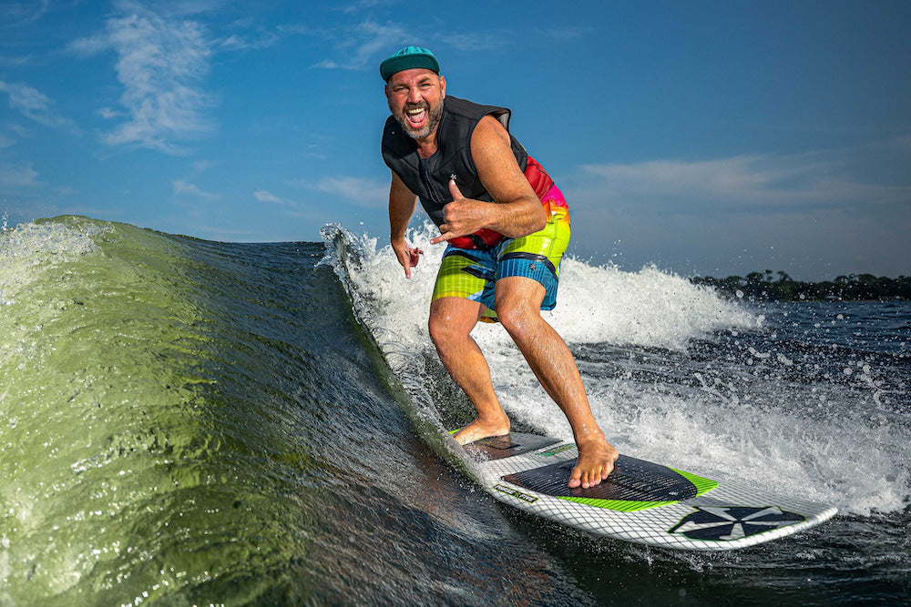 A man enjoys a wave ride with unmatched stability on a Phase 5 2025 Doctor Wakesurf Board, sporting colorful shorts and a vest beneath a clear blue sky.