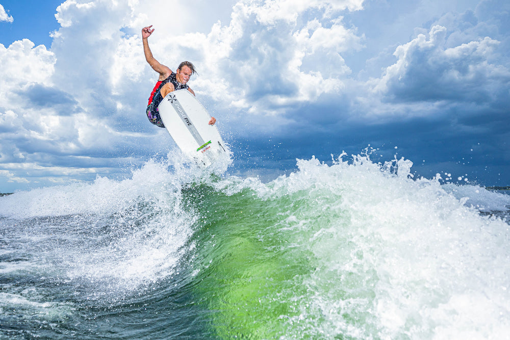 A surfer in mid-air demonstrates the ultimate stability and performs advanced tricks on a wave under a cloudy sky, riding the Phase 5 2025 Doctor Wakesurf Board.