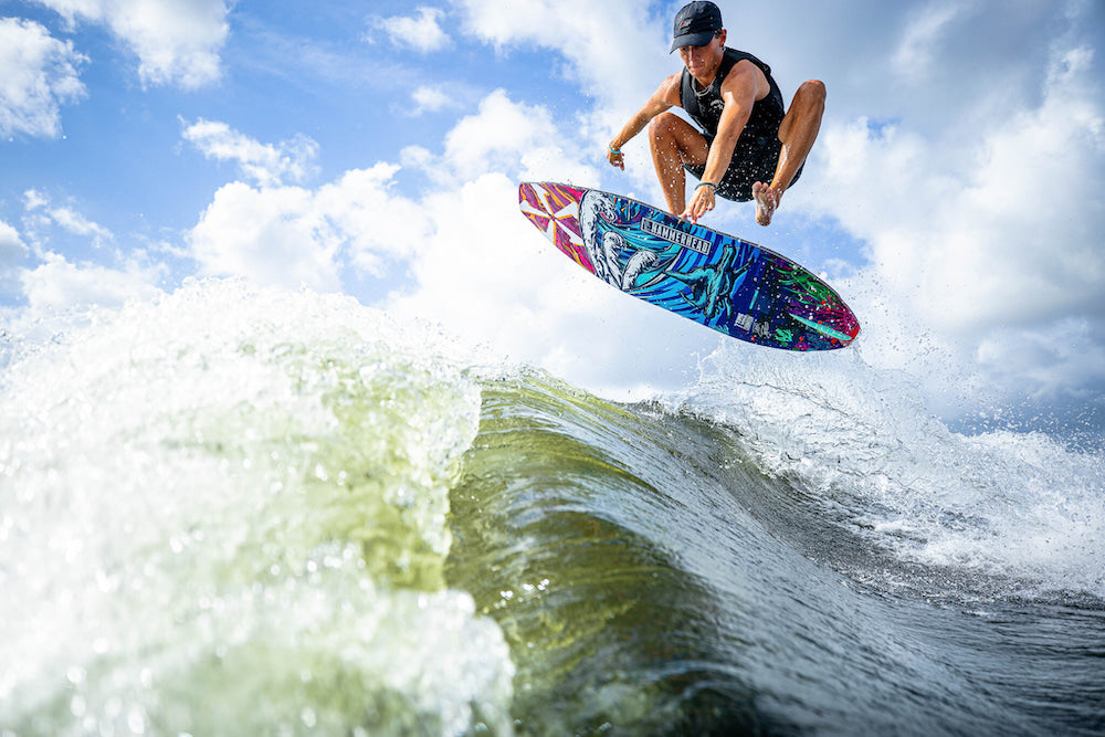 A person on a vibrant Phase 5 2025 Hammerhead v3 Wakesurf Board executes a high jump over a wave, with a cloudy sky as the backdrop.