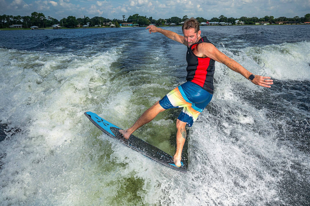 A person in colorful swimwear is surfing on a Phase 5 2025 Hammerhead v3 Wakesurf Board, expertly navigating the wake. This blue board is equipped with Hybrid Carbon™ technology, boosting performance. In the backdrop, houses and trees are seen under a partly cloudy sky, seamlessly integrating into the lively scene.