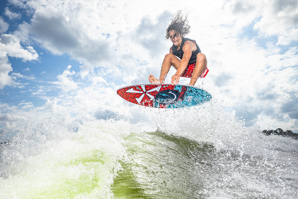 A wakesurfer on a brightly patterned wakeboard performs a jump over waves under a cloudy sky, showcasing the agility of the Phase 5 2025 Key Jett Shreds Wakesurf Board with its hybrid carbon design.