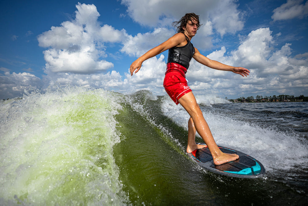 On a sunny day, someone adeptly rides a wave with their Phase 5 2025 Key Jett Shreds Wakesurf Board, while the cloudy skies in the background add drama to the scene.