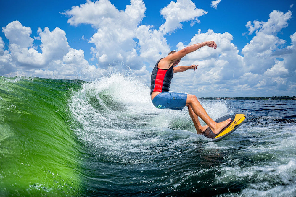 Surfing a green wave on a bright day, the rider showcases impeccable skill while wearing a wetsuit vest and shorts. The sleek Phase 5 2025 MVP Wakesurf Board from Phase 5 enhances performance, gliding effortlessly with its Hybrid Carbon technology for an exhilarating ride.