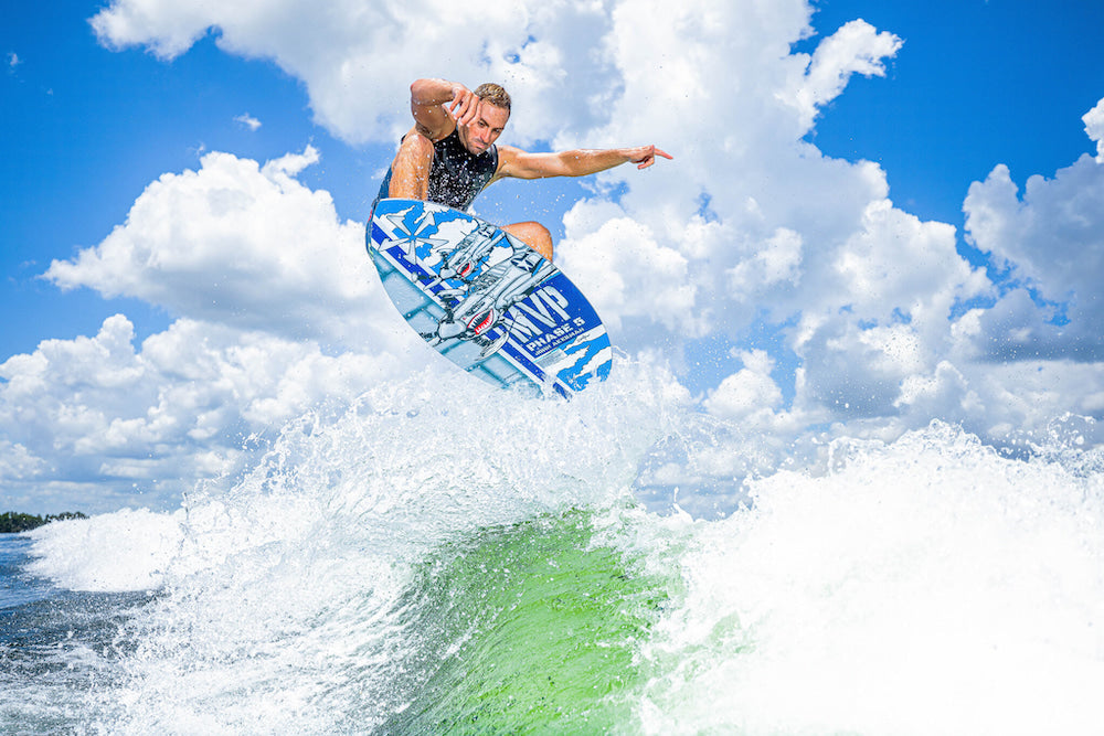 A person is surfing on a wave, skillfully performing an aerial maneuver with the Phase 5 2025 MVP Wakesurf Board. Framed by a clear blue sky and fluffy clouds, their Hybrid Carbon board provides exceptional stability, while Techno Traction V2 guarantees optimal grip for this impressive performance.