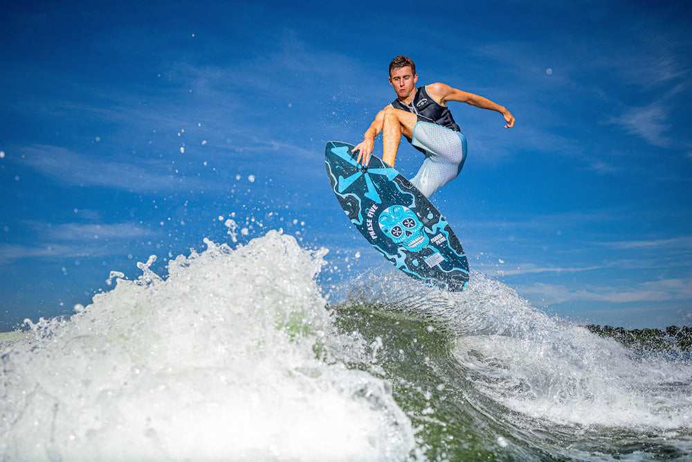 A person is executing an aerial maneuver on a wakeboard, their Phase 5 2025 Matrix Payne Pro Wakesurf Board slicing through the air with precision. The Hybrid Carbon design enhances their agility, as water splashes below and a clear blue sky spreads above.