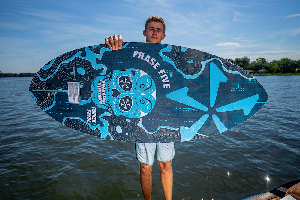 A person holds a vibrant Phase 5 2025 Matrix Payne Pro Wakesurf Board featuring a skull design, crafted with Hybrid Carbon, in front of a lake under clear skies.