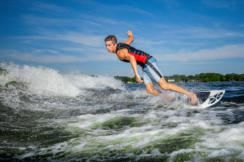 A person effortlessly glides on a wave under a clear blue sky, riding the Phase 5 2025 Phantom Wakesurf Board with precision and grace.