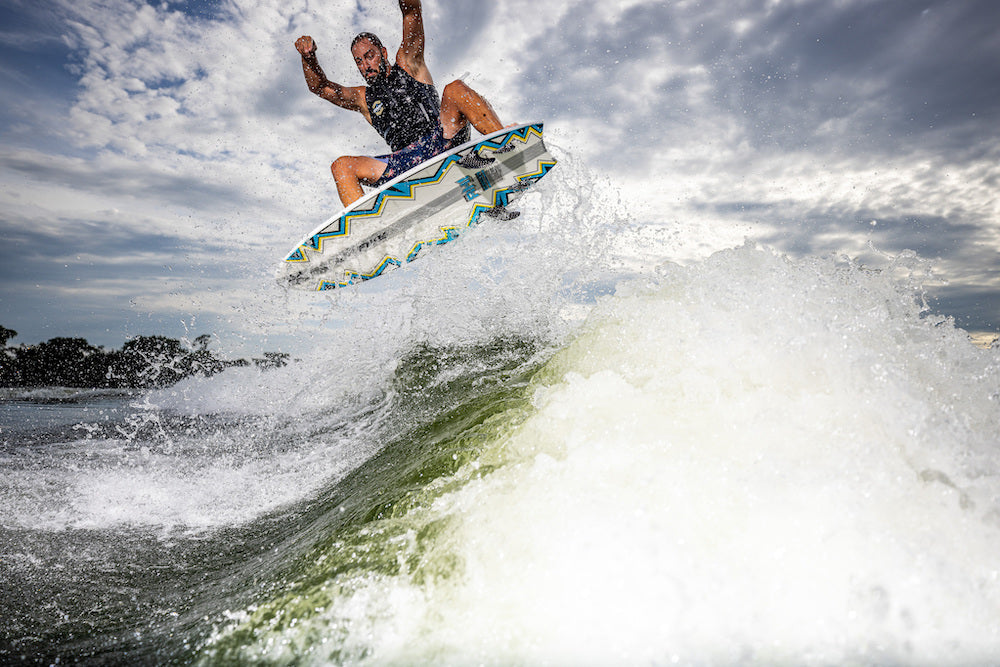 A wakesurfer in a life jacket catches air above a wave on the colorful Phase 5 Ahi Wakesurf Board 2026 by Phase 5, with water splashing around and a cloudy sky in the background.